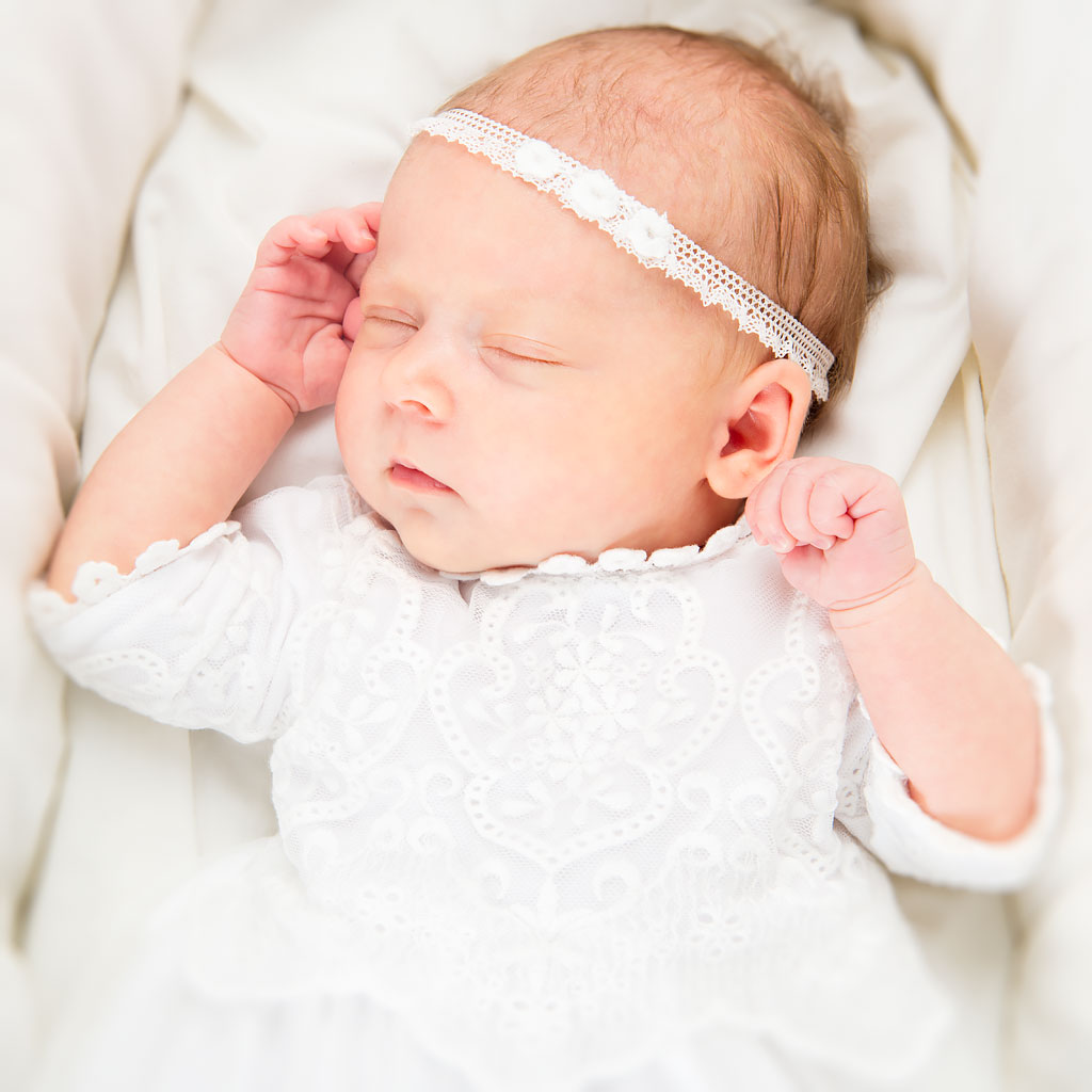 A newborn baby sleeps peacefully, wearing a delicate white baptism dress and an Eliza Lace Baby Headband in a soft, cream-colored heirloom bassinet.
