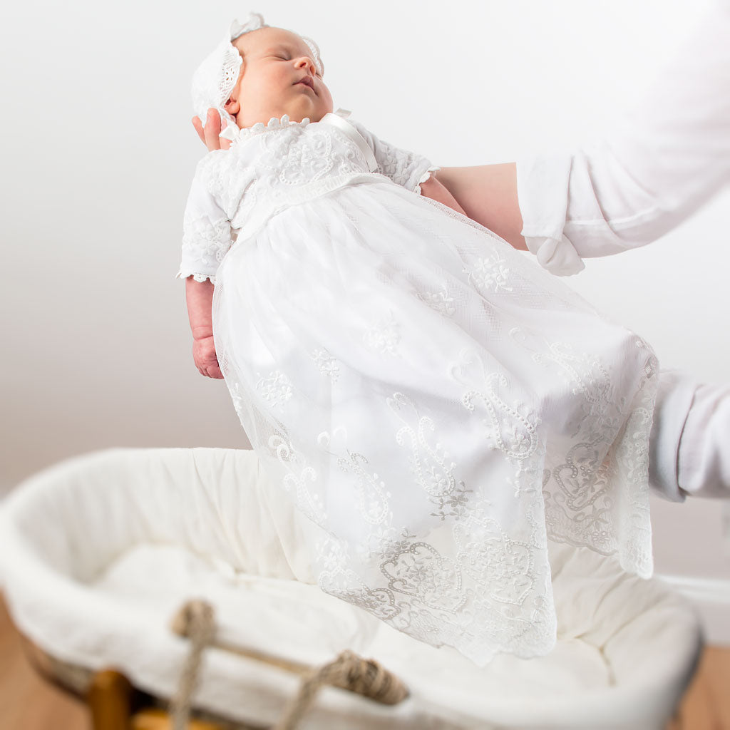 A newborn baby dressed in an Eliza Blessing Gown & Bonnet for a christening is being gently held by an adult's hands, with a soft-focus background including a vintage white bassinet.