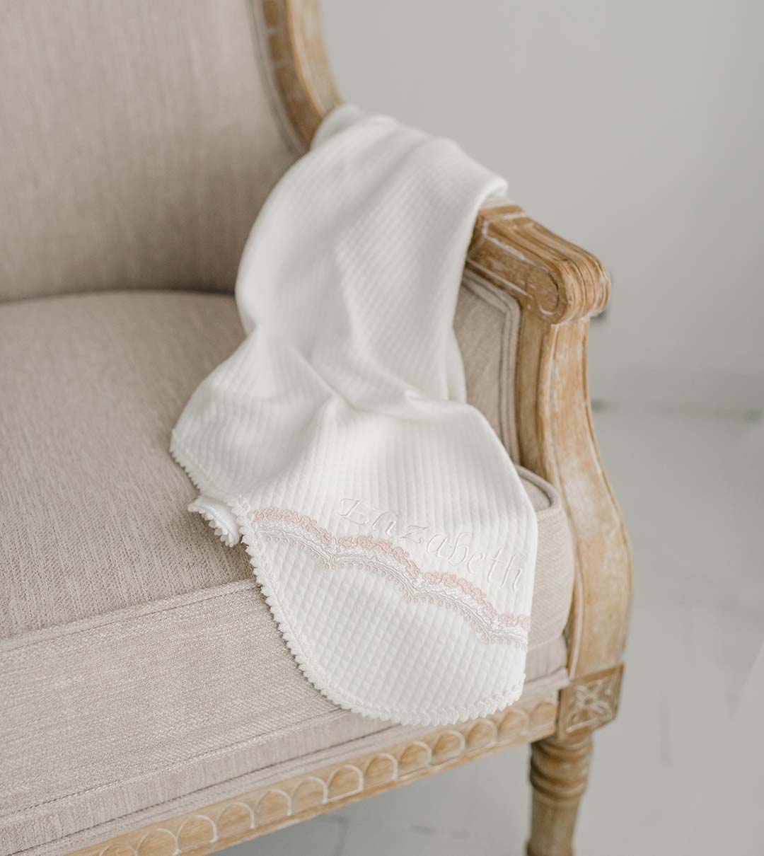 A soft white Elizabeth Personalized Blanket with pink embroidery for baptism is draped over an elegant beige vintage armchair, positioned against a plain white background.