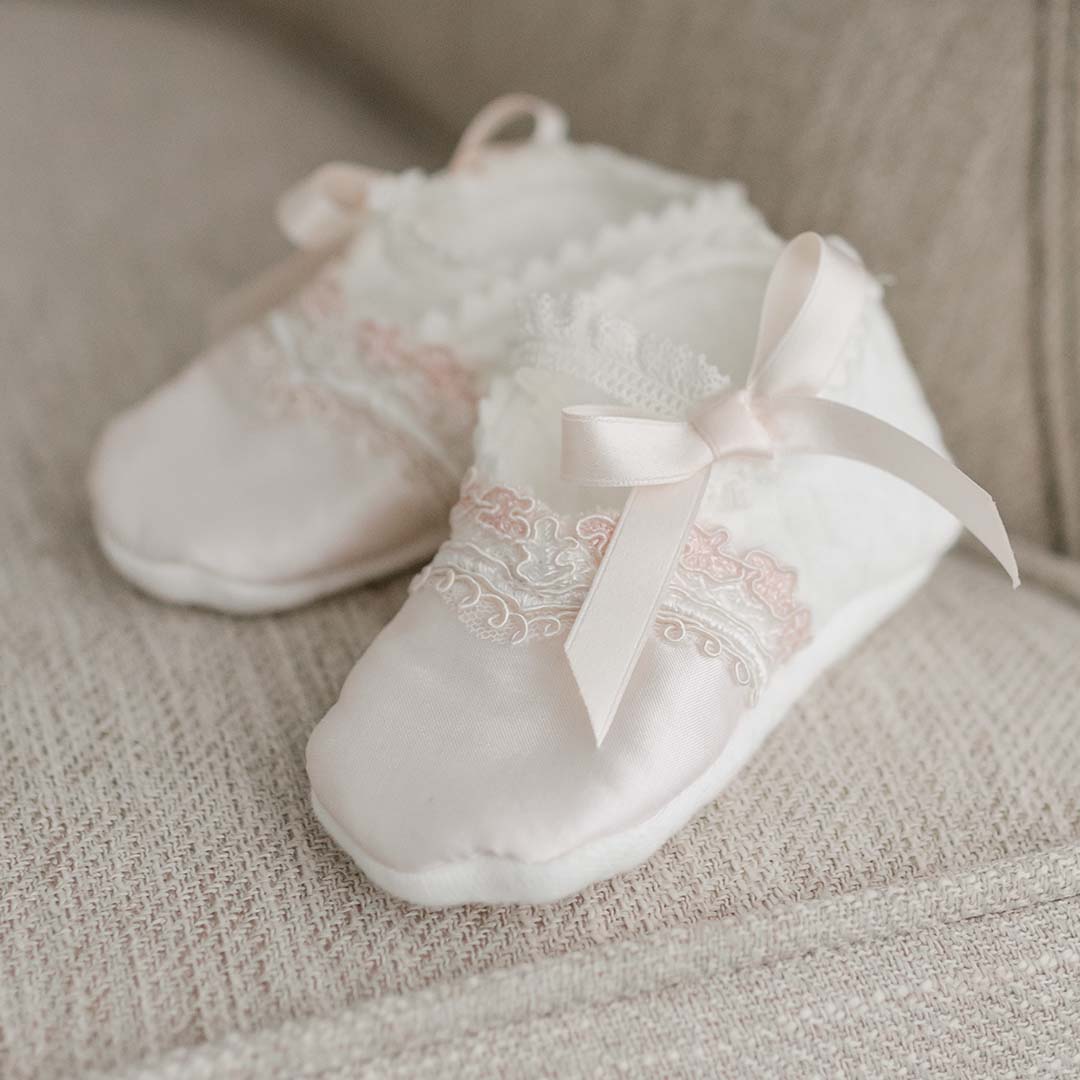 A pair of delicate white Elizabeth Booties with pink ribbons and lace detailing, resting on a soft beige fabric surface.