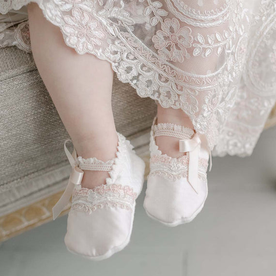 A close-up of a baby's feet wearing delicate Elizabeth Booties, possibly an heirloom, with satin ribbons, dangling in the air against a soft, pale background.