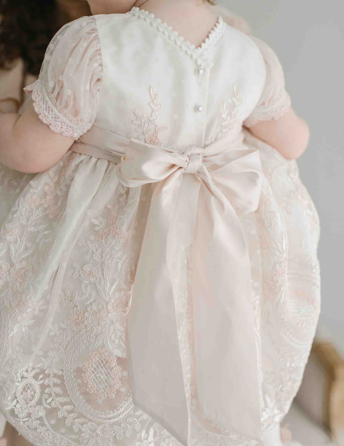 A toddler in an Elizabeth Christening Dress with detailed embroidery and a large satin bow on the back, standing indoors, showing only their back, ready for a baptism.