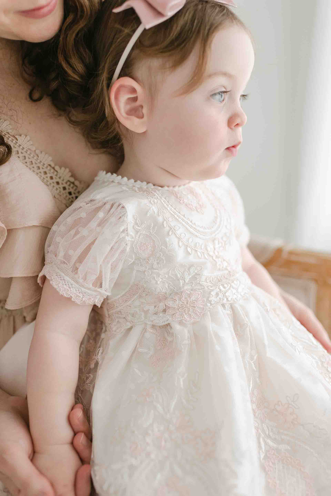 A toddler in a delicate white Elizabeth Christening Dress, held by her mother, gazes thoughtfully out of a window.