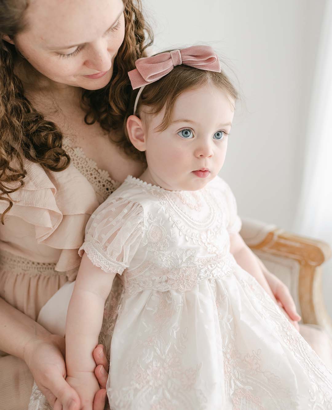 A mother gently holds her toddler, dressed in the Elizabeth Christening Dress with lace details for a christening. The child looks surprised as they sit by a window with soft natural light.