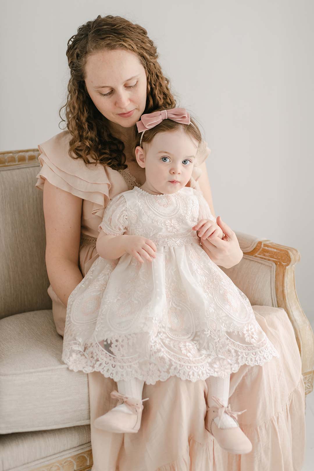 A woman with curly hair and a pink dress tenderly holds a baby girl in an Elizabeth Christening Dress on her lap, seated on an upscale light beige couch. Both are looking directly at the camera.