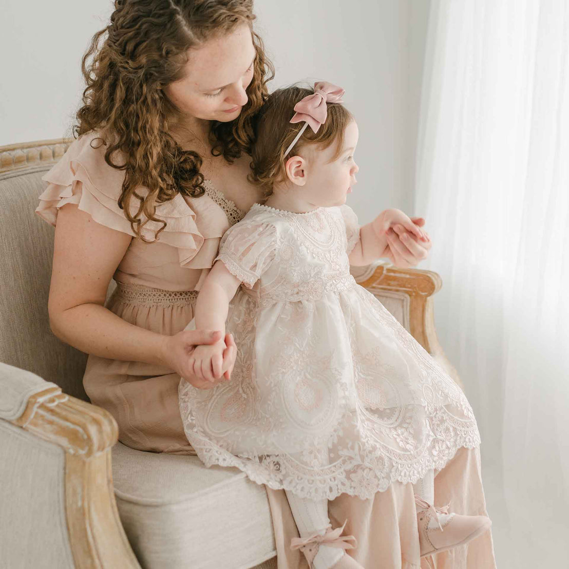 A mother with curly hair in a pink blouse and a toddler girl in an Elizabeth Christening Dress, sit together on an elegant vintage sofa in a bright room, sharing a tender moment