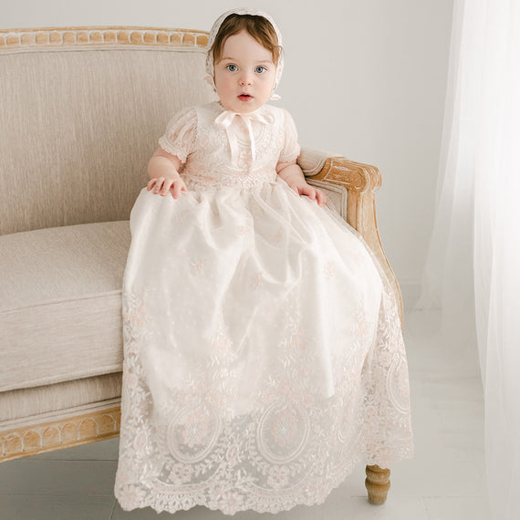 A baby girl in an Elizabeth Christening Gown & Bonnet sits on a beige vintage sofa, looking directly at the camera with a curious expression. The room has a soft, neutral color scheme.