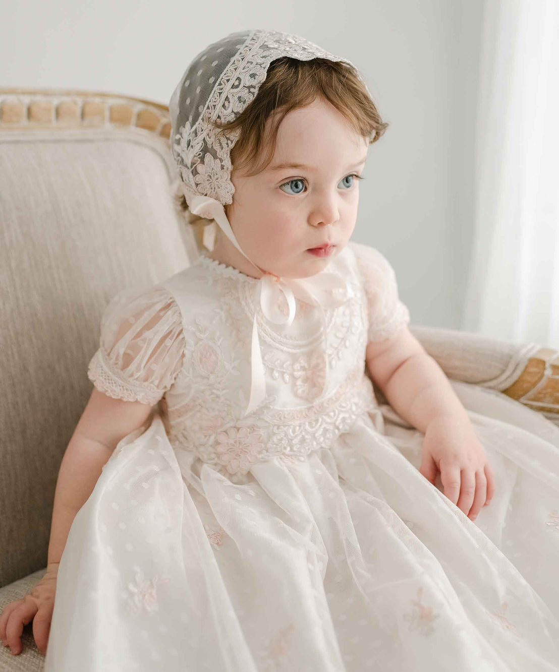 A baby girl with wide blue eyes sits on a beige sofa, wearing the Elizabeth Christening Gown & Bonnet, looking slightly off-camera.