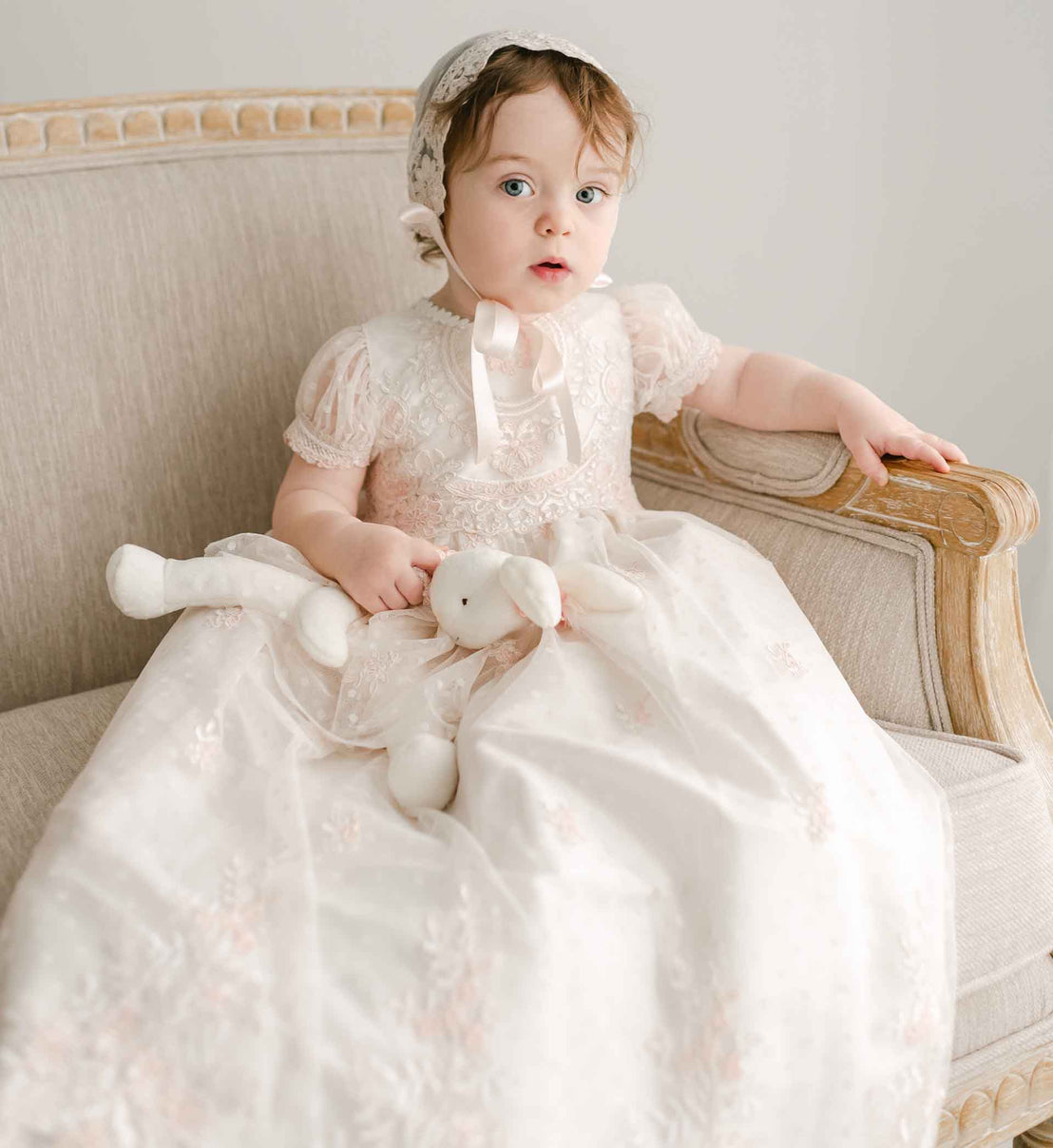 A baby girl in the Elizabeth Christening Gown & Bonnet sits on a beige sofa, holding the Elizabeth Silly Bunny Buddy, a white stuffed bunny.