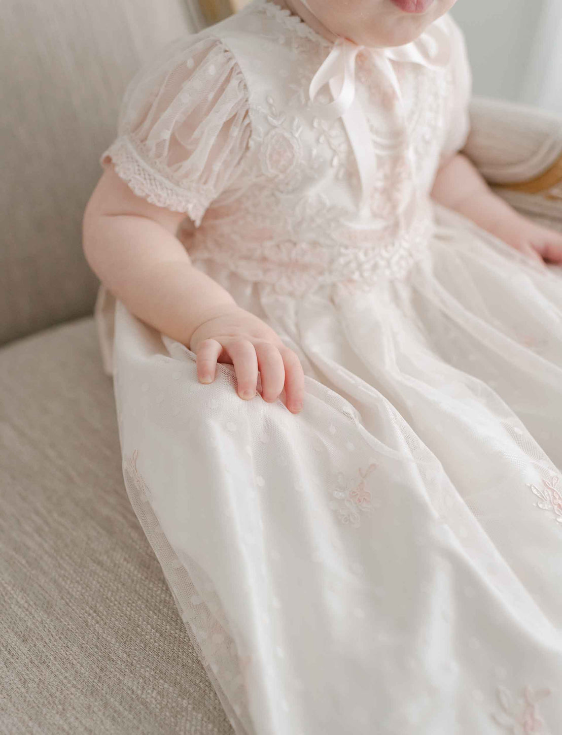 A close-up of a baby wearing the Elizabeth Christening Gown, focusing on the detailed fabric and the child's small hand resting on the gown.