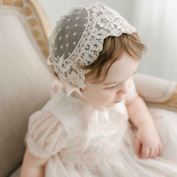 A baby wearing the Elizabeth Lace Bonnet and dress for a christening, sitting on a vintage sofa, looking down thoughtfully in a soft and light-colored setting.