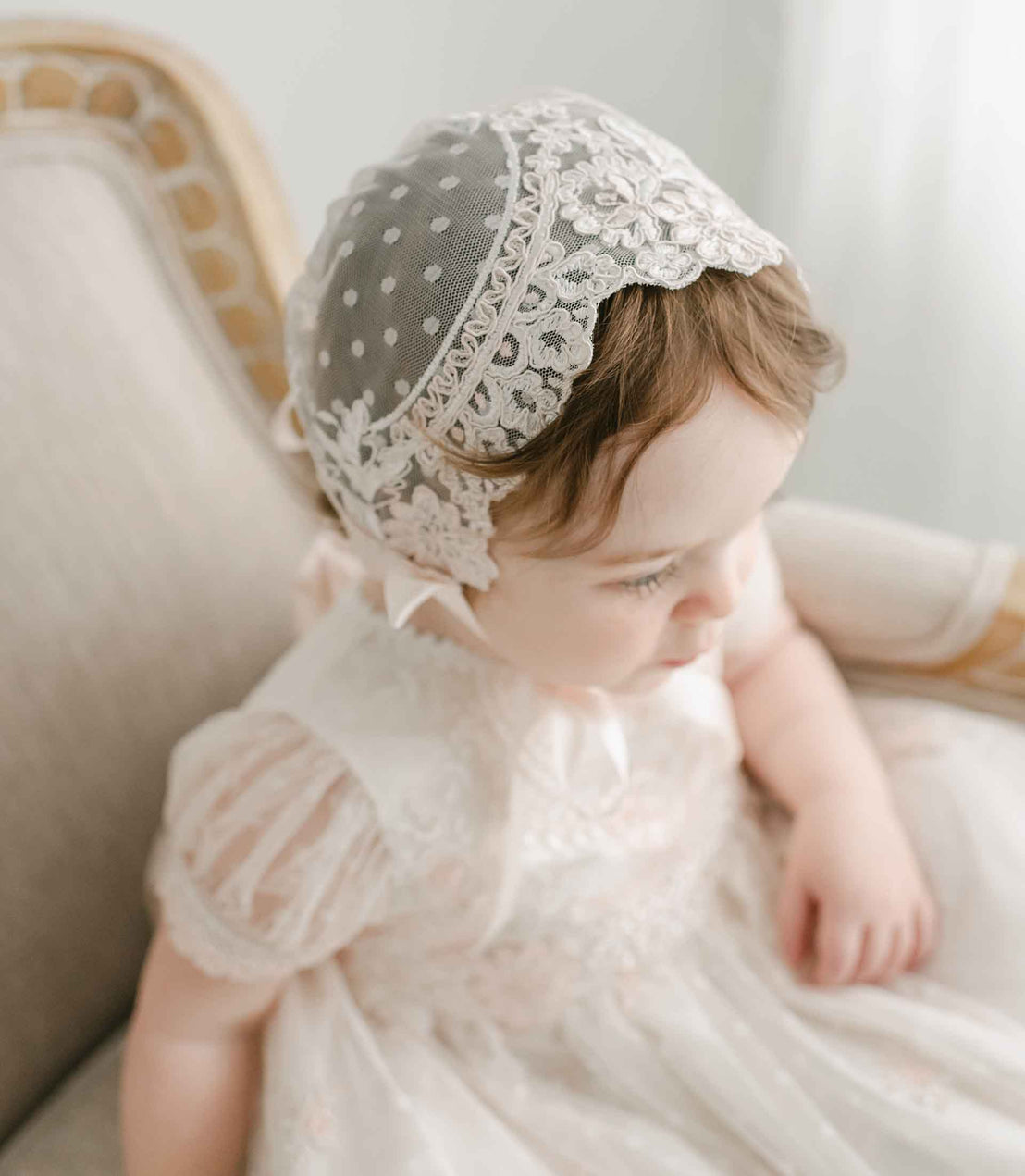 A baby girl wearing the Elizabeth Christening Gown & Bonnet looks down thoughtfully while sitting on an elegant vintage chair.