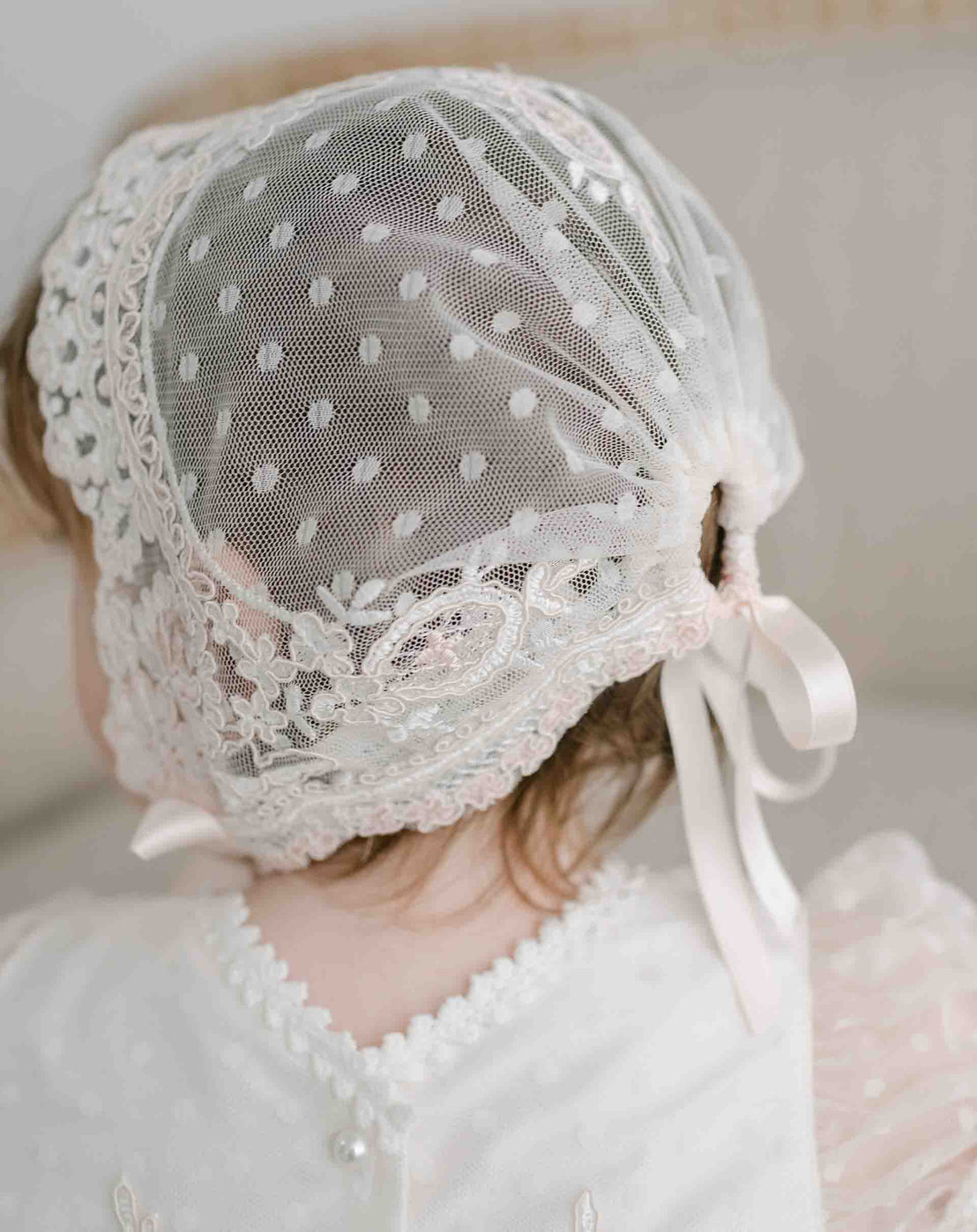 A baby girl wearing the Elizabeth Christening Gown Bonnet viewed from behind. The bonnet features a polka dot lace pattern, secured with a soft pink ribbon. The bonnet is sheer and embroidered with floral details.
