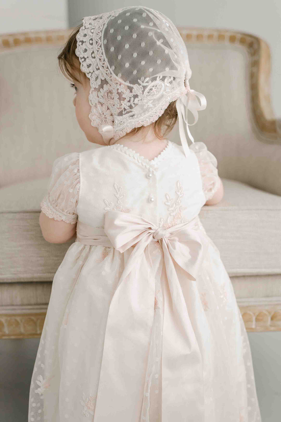 A baby girl in an Elizabeth Christening Gown & Bonnet stands facing away from the camera, focusing on something off-camera, in a softly lit room.