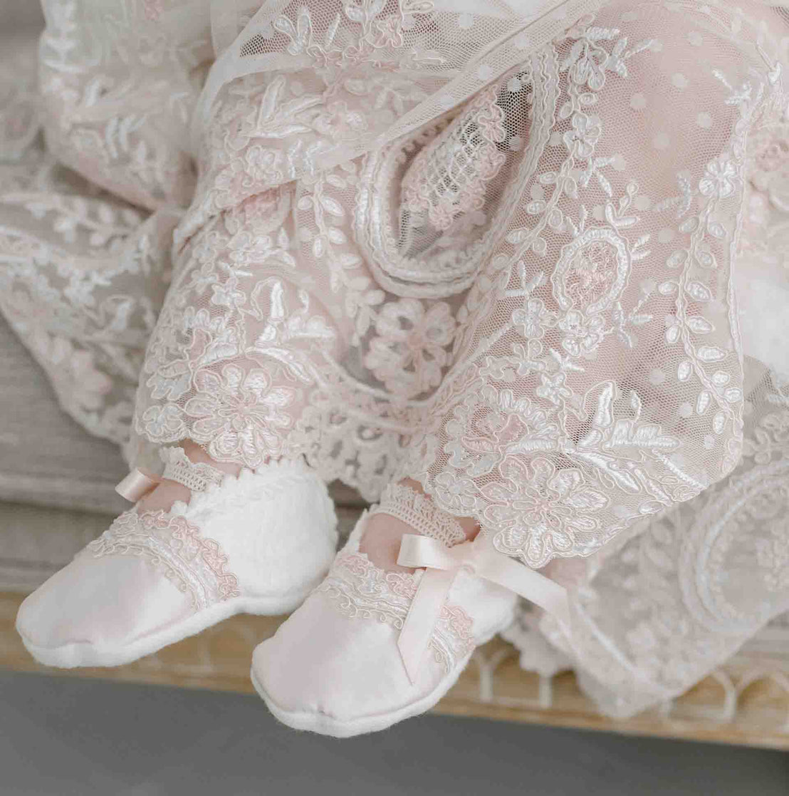 Close up detail of the Elizabeth Christening Gown's skirt, as worn by a baby girl, featuring the intricately detailed pink and ivory dotted mesh embroidered lace with cording. The baby girl is also wearing the Elizabeth Booties made from ivory quilted cotton and detailed with ivory and pink edge lace.