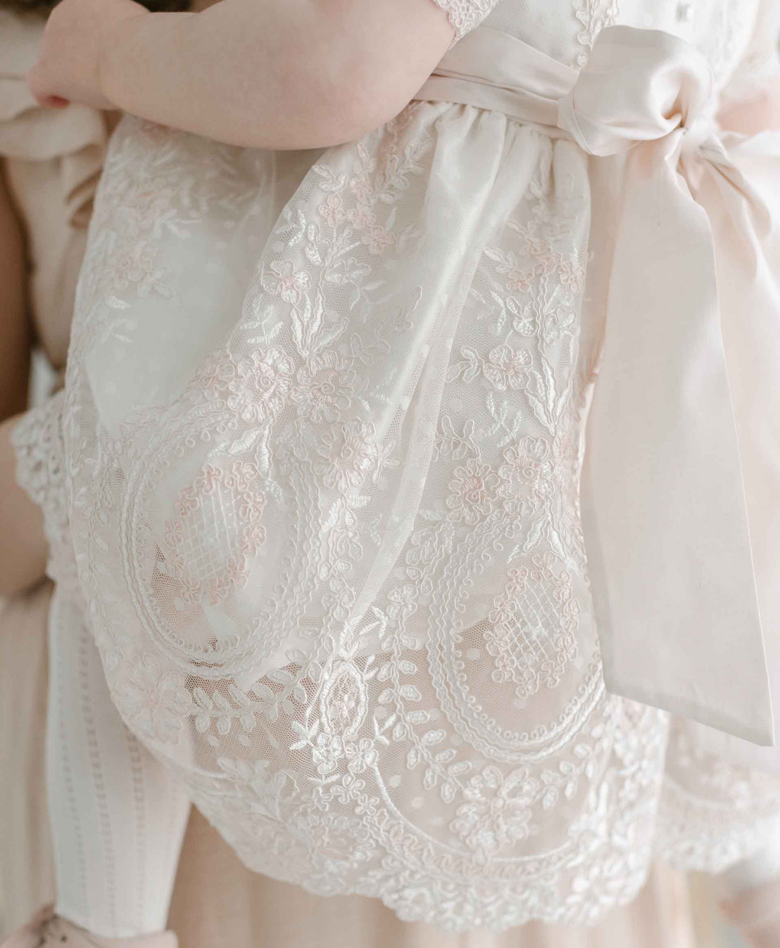 A close-up of a person holding the delicate, lace-adorned fabric of a Elizabeth Christening Dress, featuring intricate embroidery and a bow.