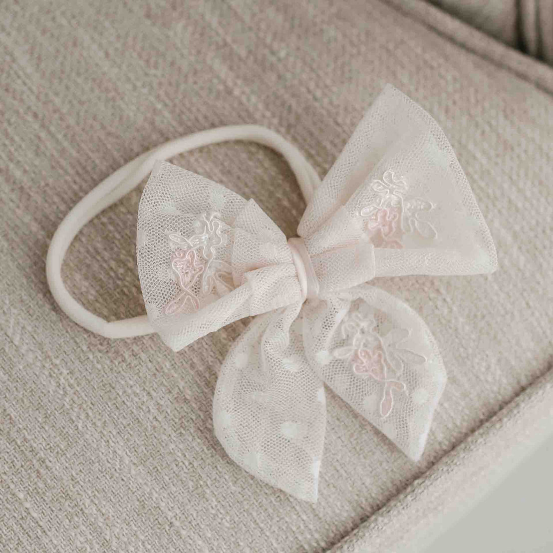 A delicate, beige Elizabeth Lace Bow Headband topped with a large, decorative bow featuring intricate white floral embroidery, ideal for christening and displayed on a soft, textured cushion.