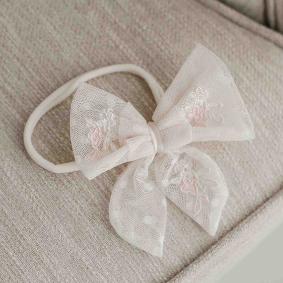 A delicate, beige Elizabeth Lace Bow Headband topped with a large, decorative bow featuring intricate white floral embroidery, ideal for christening and displayed on a soft, textured cushion.