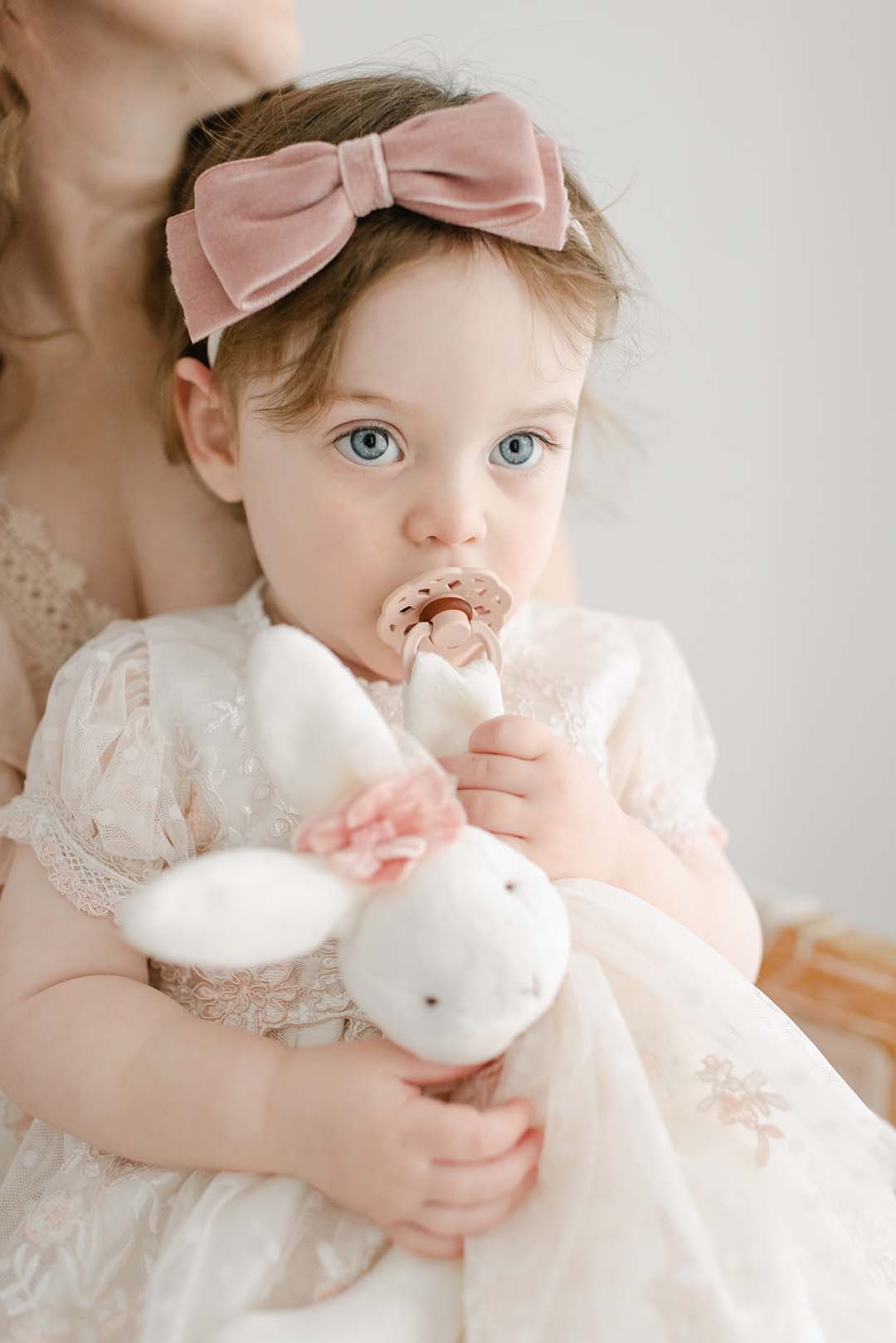A toddler girl with blue eyes holds a white Elizabeth Silly Bunny Buddy and sucks on a pacifier. She wears an heirloom lace dress and a pink bow headband, partially held by an adult whose face is.