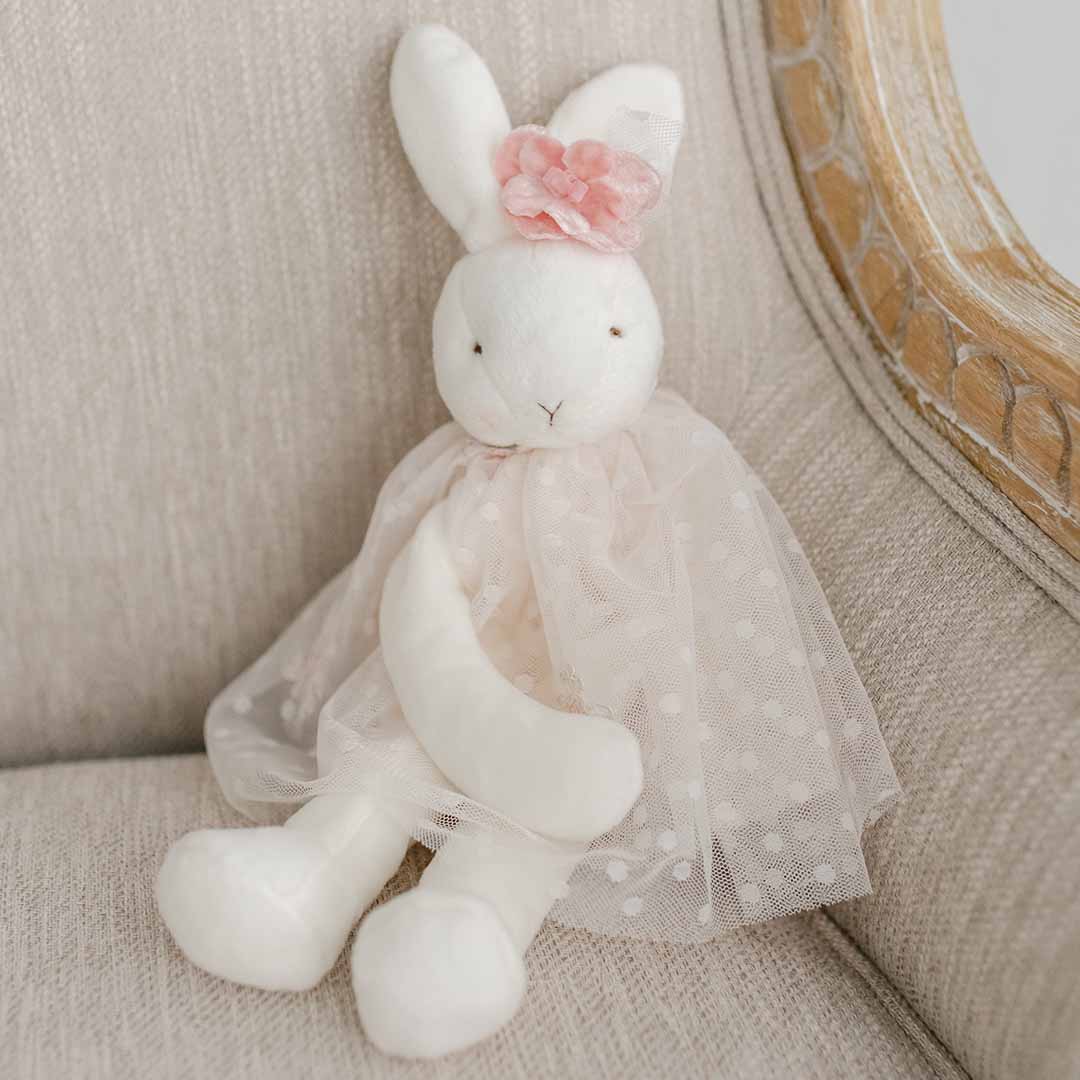 A vintage Elizabeth Silly Bunny Buddy | Pacifier Holder toy with a pink flower headband, wearing a sheer tulle dress for baptism, sits on a textured beige armchair.