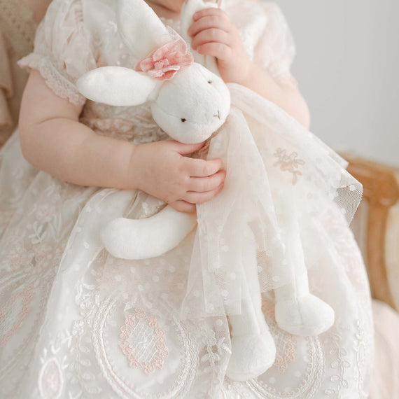 A toddler in an elegant heirloom lace dress holds a plush Elizabeth Silly Bunny Buddy during a christening. The focus is on the child's hands, the bunny, and the intricate dress details.