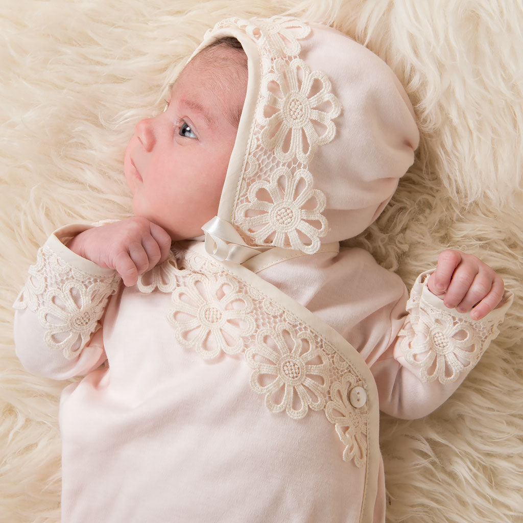 A newborn baby in a pink Hannah Knot Gown & Bonnet with lace detailing and lying on a soft, fluffy cream-colored blanket.