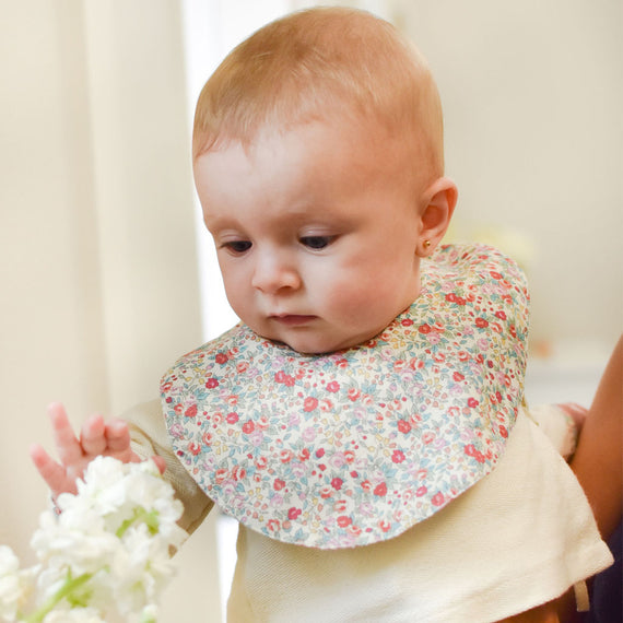 A baby with a Petite Fleur Bib reaches out to touch white flowers, showing curiosity and focus, in a brightly lit room.