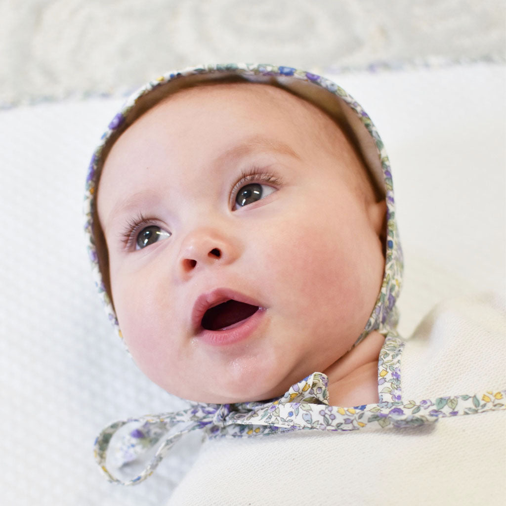 Close-up of a baby with wide eyes wearing a Petite Fleur Bonnet, lying on a white background, looking upward with a surprised expression.