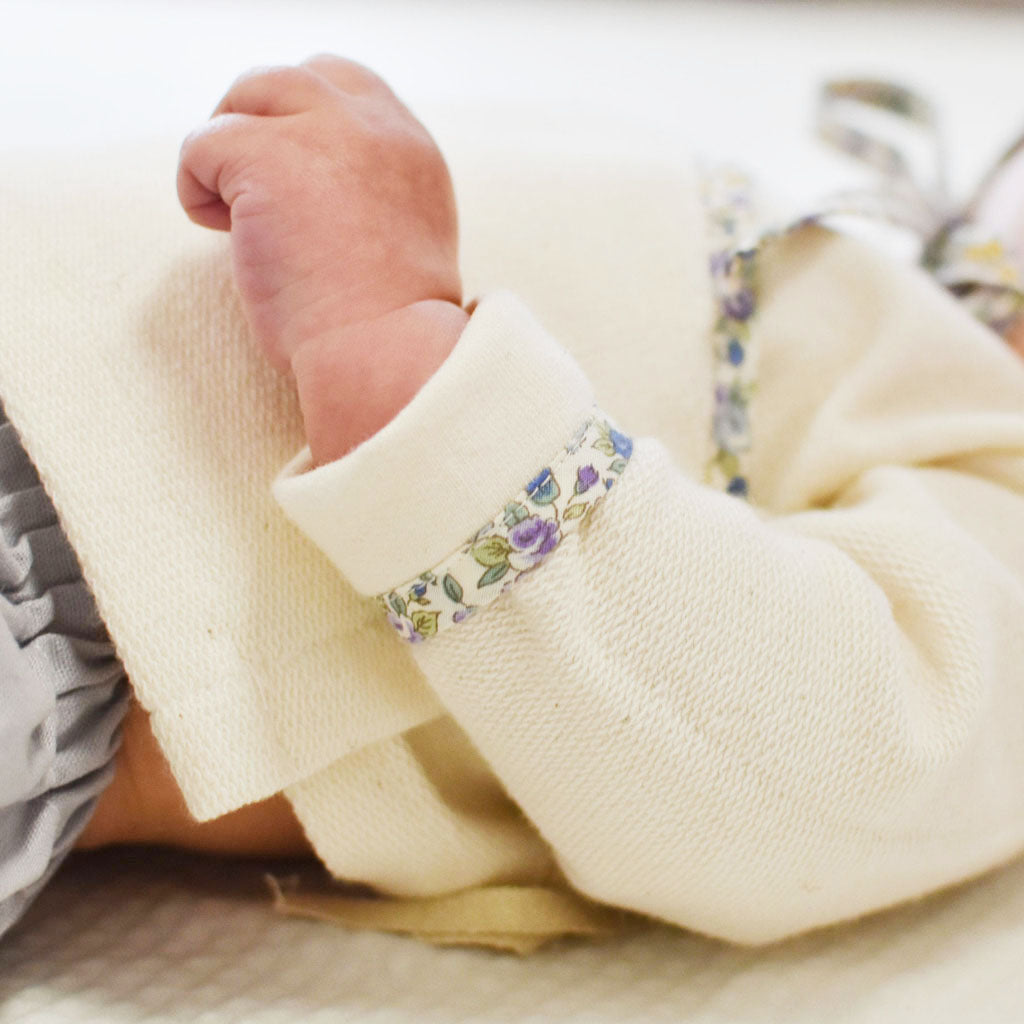 A close-up of a newborn's tiny fist emerging from a Petite Fleur Wrap Top & Pants in cream color, with floral trim on the cuff, set against a soft, off-white background.