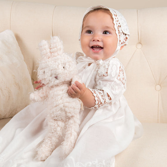 Baby girl smiling a plush bunny doll while wearing a cotton baptism gown and bonnet. Bunny doll is part of the matching Adeline collection. 