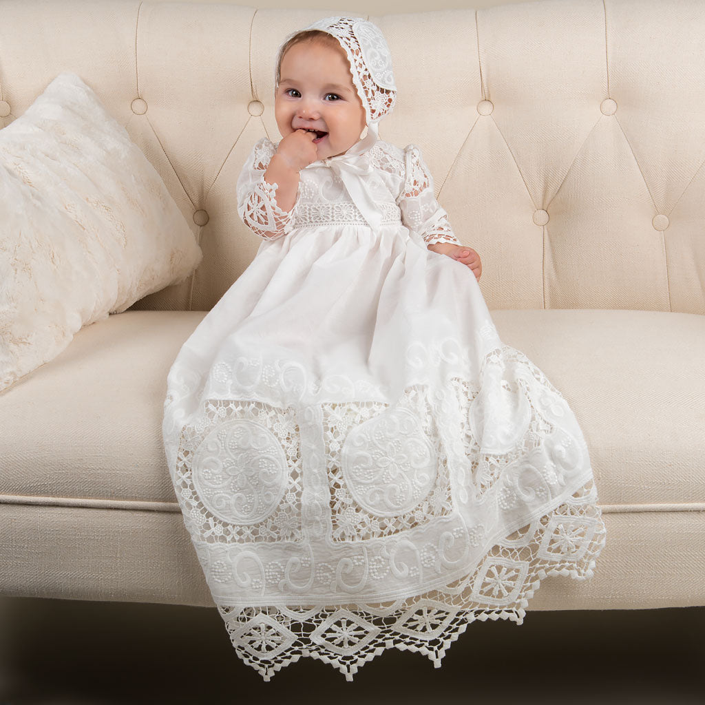 Baby girl smiling with hand in her mouth wearing a beautiful white cotton baptism dress. Part of the Adeline Lace christening collection.
