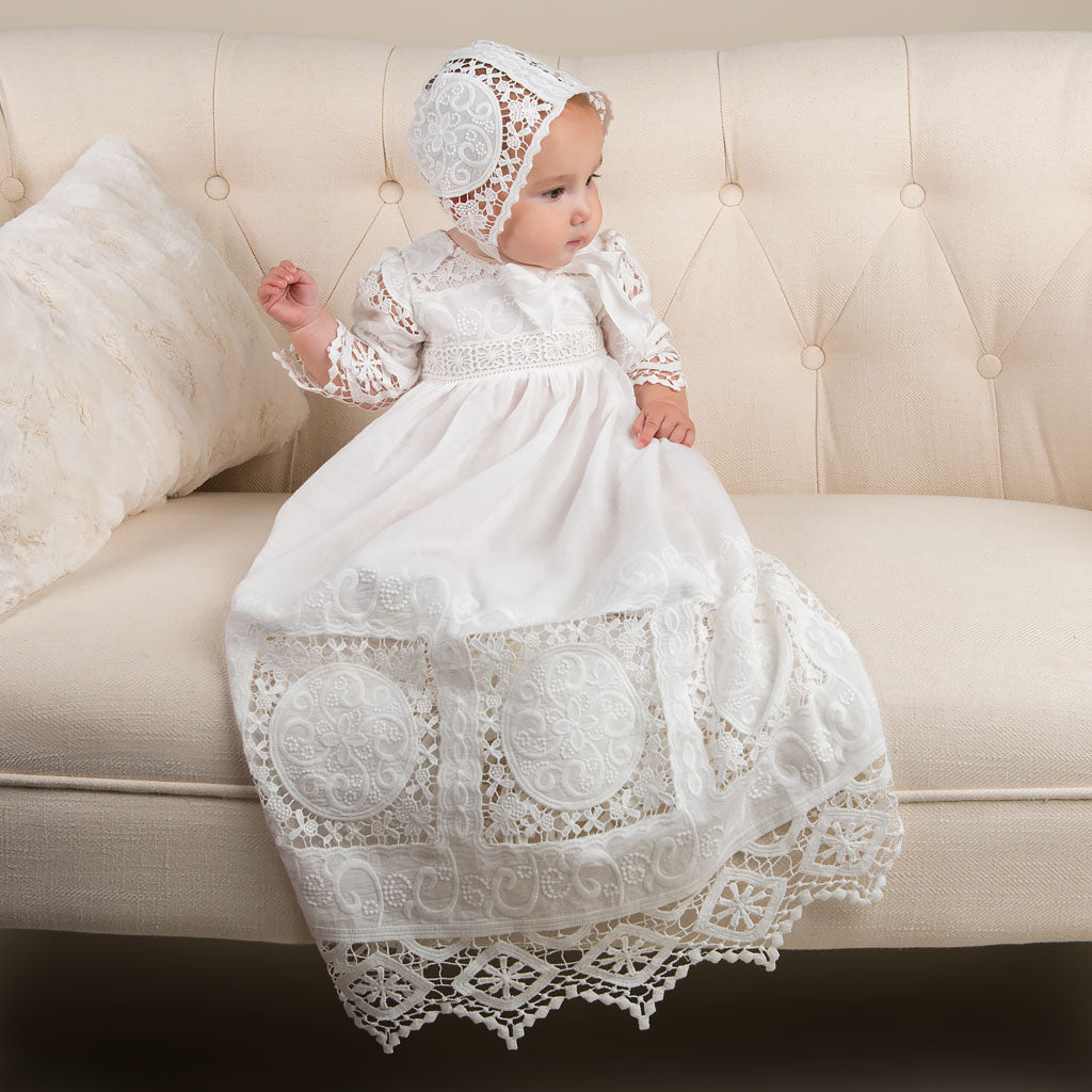 Baby girl wearing the Adeline lace christening gown made of white cotton. Pictured on a beige couch.