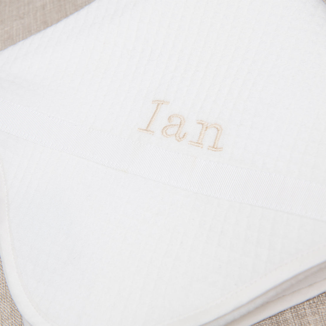 Close-up view of a designer white baby blanket with a gray stripe and the name "Ian" embroidered in beige thread.