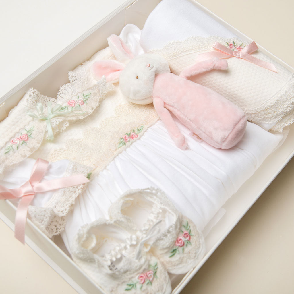 A Baby Beau & Belle Ivory Gift Box with a magnetic closure containing a plush bunny, knitted baby clothes, and lace-trimmed accessories arranged neatly, all in soft pastel colors.