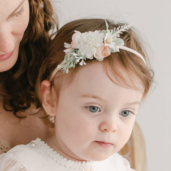 A toddler with blue eyes, wearing a white Elizabeth Flower Headband heirloom dress, looks to the side while being held by a woman with curly hair, visible only partially and out of focus.