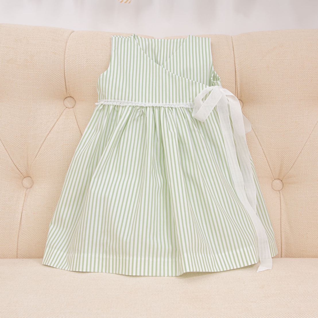 The sage green Thea Wrap Dress displayed on a beige sofa. The cotton ribbon bow on the waist is featured in the photo.