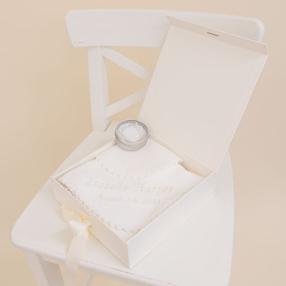 A Baby Beau & Belle Baby Girl Personalized Gift Set, including a baptism candle and a personalized white cotton bib embroidered with the name "Isabella Harper" and the date "August 14, 2021," displayed in an open white box on a white chair.