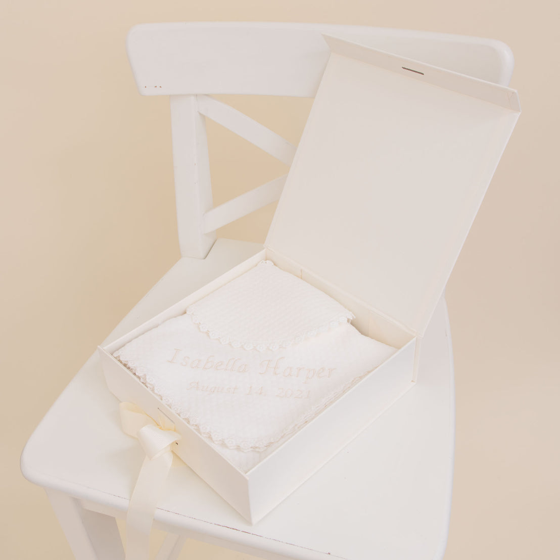 A white chair with a Baby Beau & Belle Baby Girl Personalized Gift Set on the seat, containing a quilted cotton bib embroidered with "Isabella Harper August 14, 2021" in elegant script.