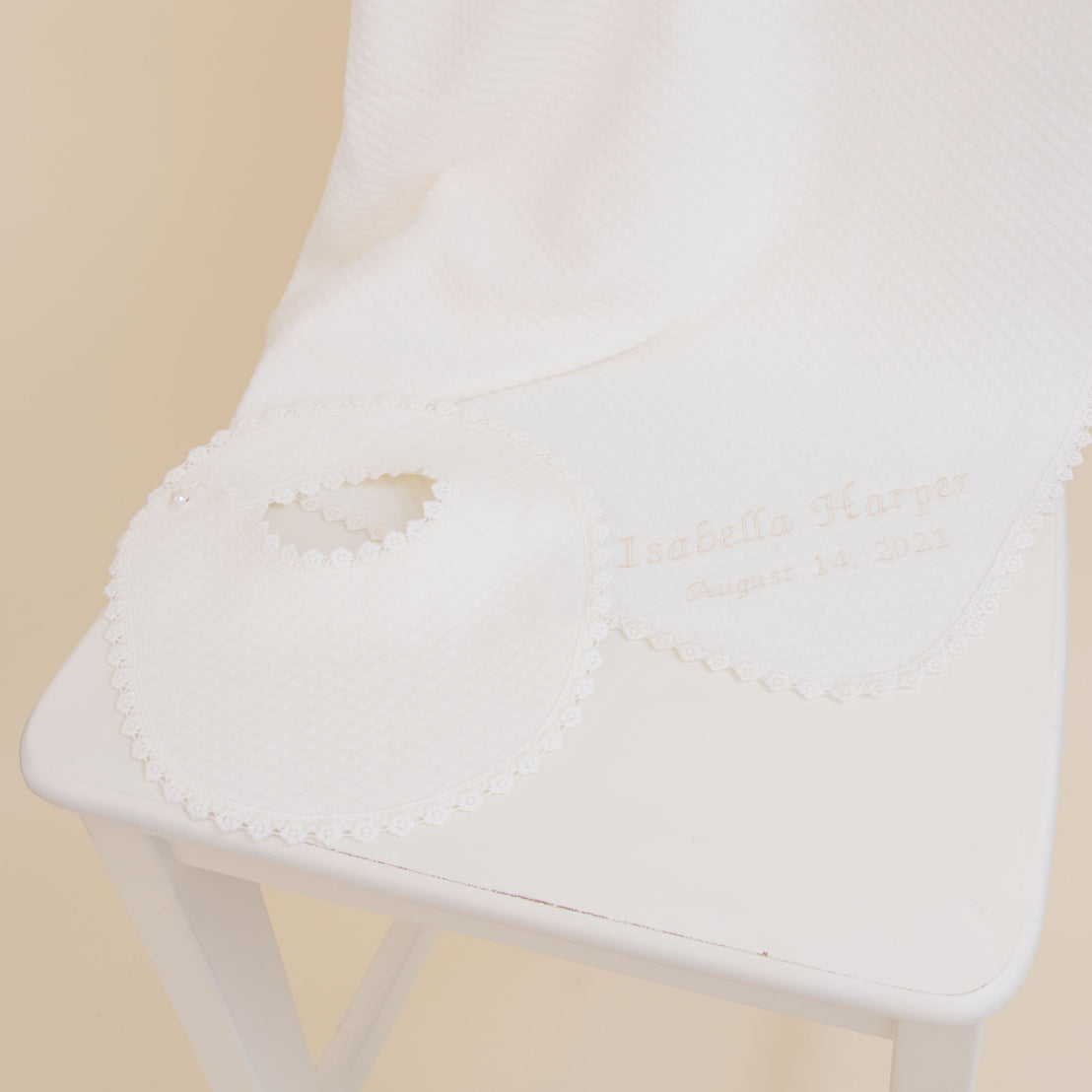 A white Baby Beau & Belle baby blanket and bib personalized with the name "Isabella Harper" and the date "August 14, 2021," laid on a white chair with a beige background. The cotton bib