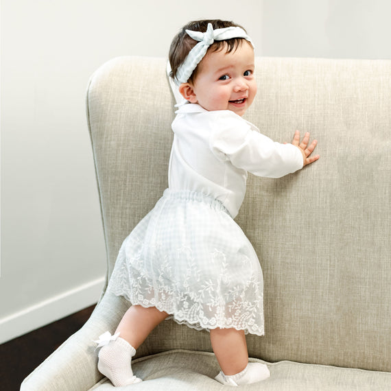 A smiling newborn girl stands by holding onto a gray couch, dressed in the Isla Bubble Skirt Set and matching Tie Headband. 