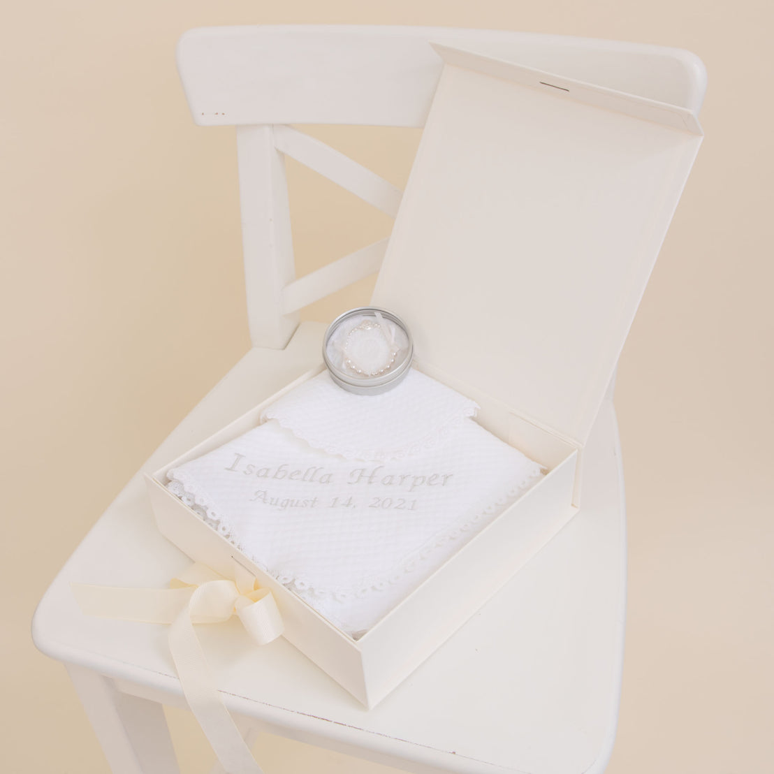 A white memory box on a chair, containing a Baby Beau & Belle Baby Girl Personalized Gift Set—a cushion with a silver rattle and an embroidered baby blanket reading "Isabella Harper, August 15, 2021.