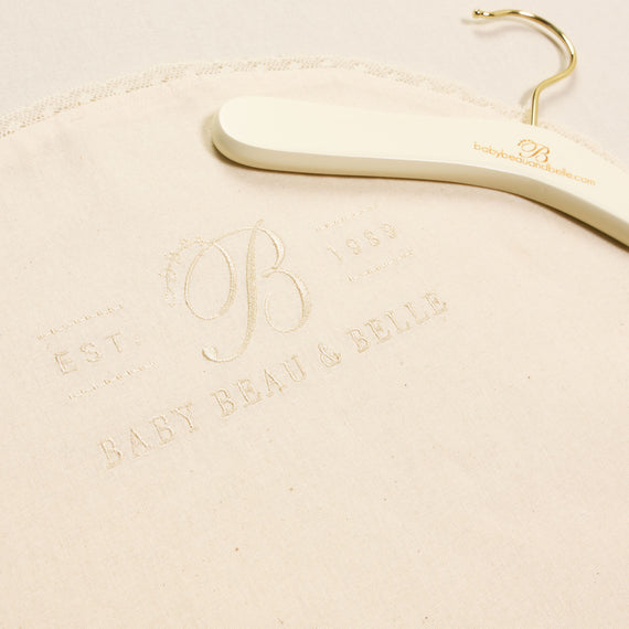 A close-up photo of a Muslin Keepsake Garment Bag with an embroidered heirloom logo "baby beau & belle" on a vintage textured fabric with an elegant hanger and the year "1989" established date.