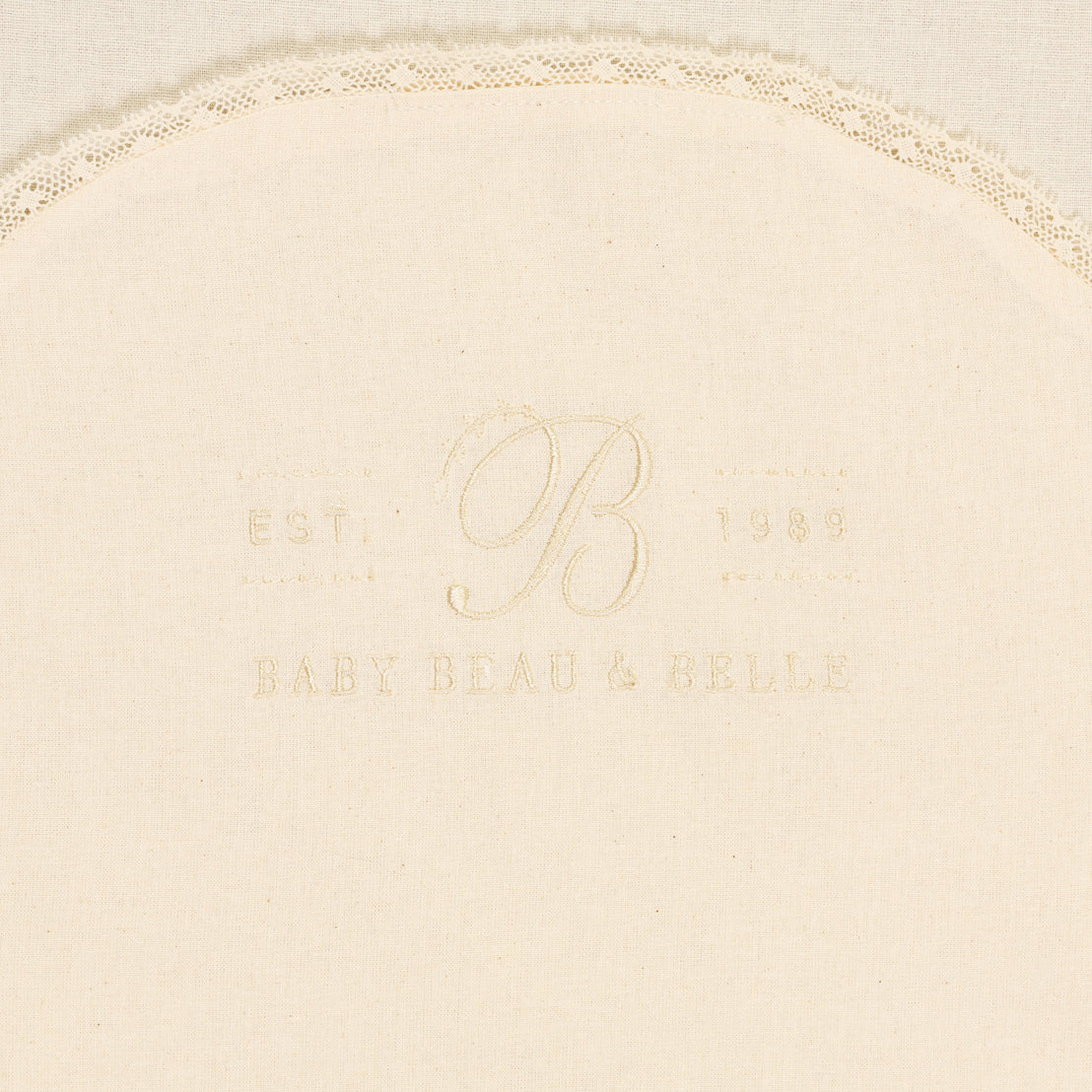 Close-up of an heirloom embroidered logo that reads "est. b 1989," with "baby beau & belle" below in faint stitching on ivory fabric with christening lace trim at the top Muslin Keepsake Garment Bag