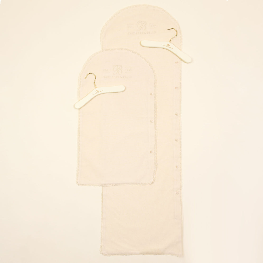 Two beige Muslin Keepsake Garment Bag covers with white hangers on a light background, featuring visible button details and labels at the top, ideal for baptism or christening attire.