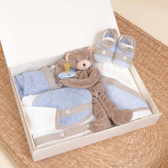 An Austin Newborn Gift Set containing a plush teddy bear, baby booties, a pacifier, and folded baby clothes, placed on a wicker surface.