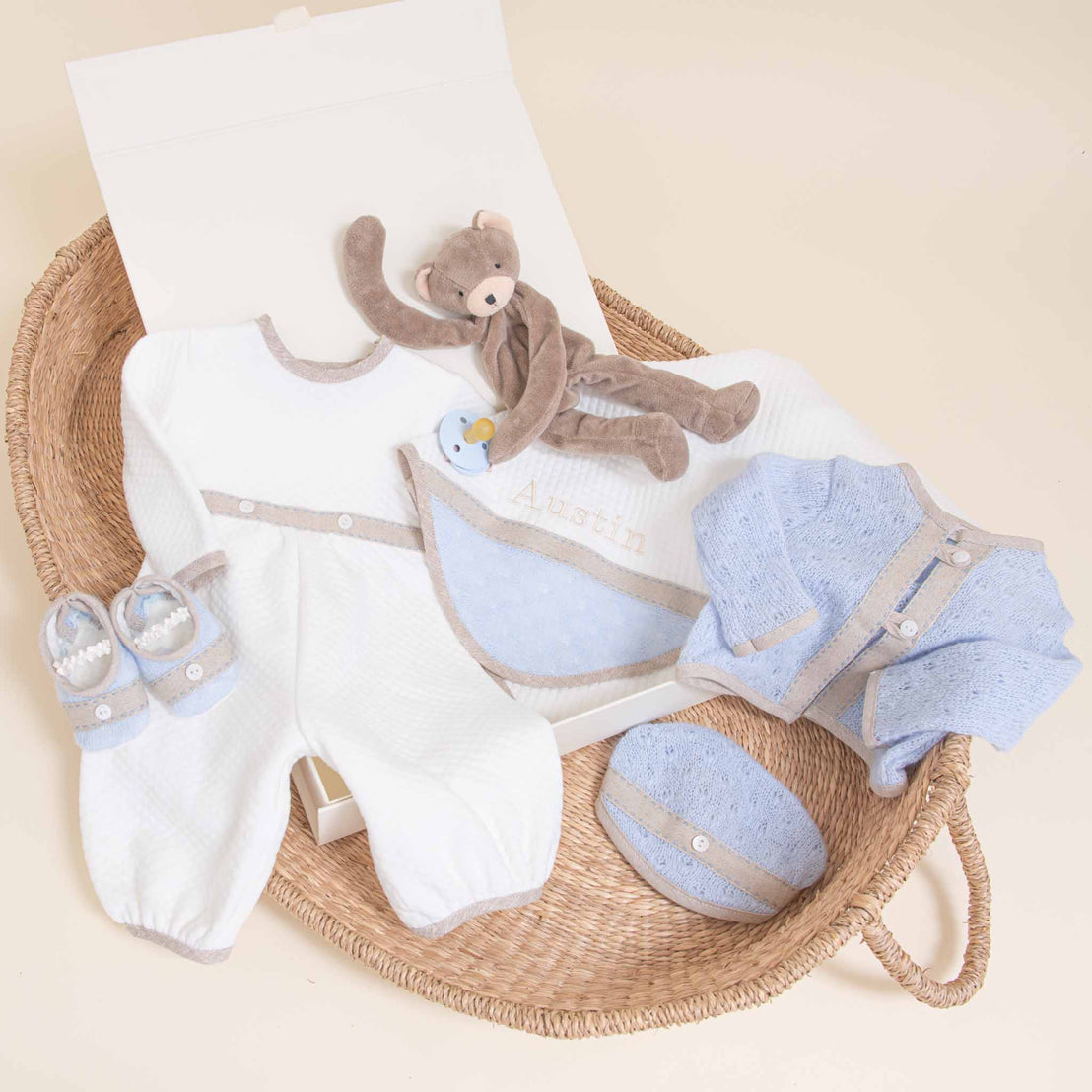 A collection of Austin Newborn Gift Sets in a wicker basket, including a white onesie, a blue knit cardigan, matching booties, and a plush bear toy.