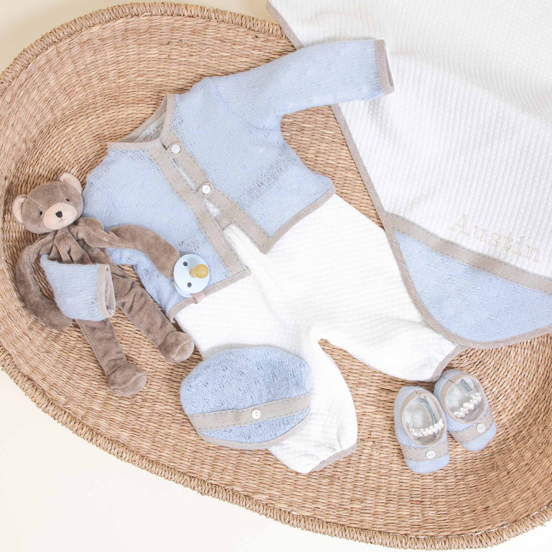 A collection of Austin Newborn Gift Set items laid out on a wicker mat, including an heirloom blue romper, a white personalized blanket with a name, a teddy bear, bib, and socks.