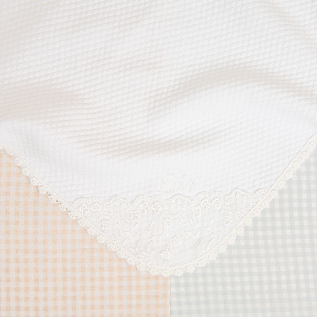 Close-up of a traditional white embroidered Isla Personalized Blanket with a floral pattern and the name "ella" embroidered, designed as an heirloom for a newborn, against a backdrop of checkered fabric in soft.