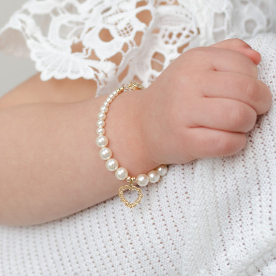 A close-up image of a baby's wrist adorned with a Cream Luster Pearl Bracelet with 14k Gold Heart Charm, featuring a gold clasp and a pendant shaped like a heart. This charming piece of baby jewelry makes an excellent baby gift.