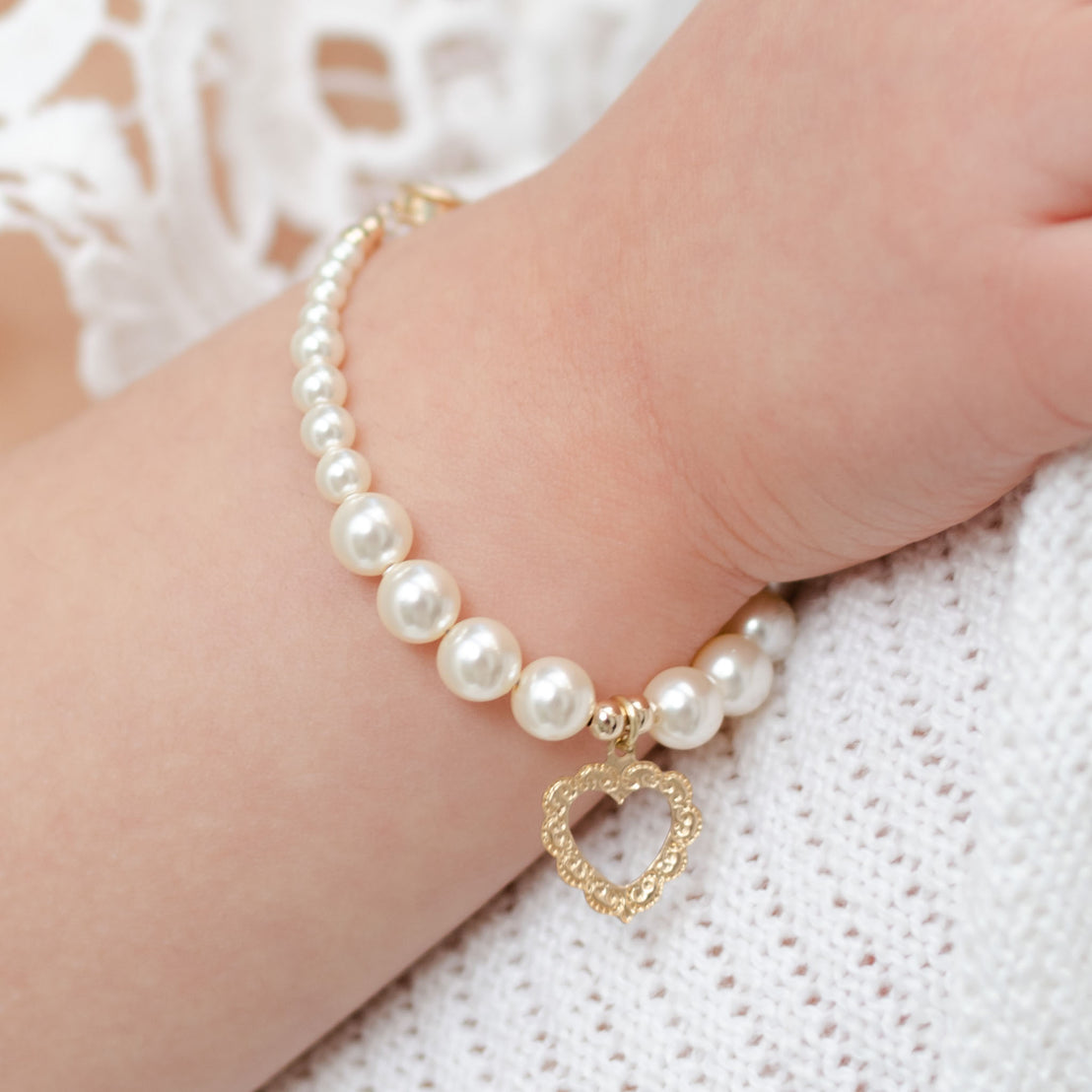 Close-up of a woman's wrist wearing a Cream Luster Pearl Bracelet with 14k Gold Heart Charm, against a soft white lace and fabric background at a baby shower.